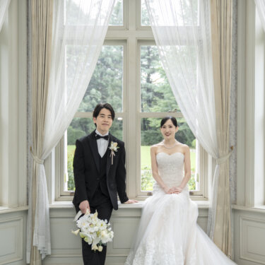 白いカーテンの出窓に並んで腰かける、ブーケを持った新郎とウェディングドレスの花嫁。旧古河邸の明るい洋館で撮るフォトウェディング