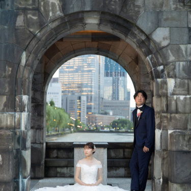 石造りのアーチの下で、ドレスを広げて座る花嫁と柱にもたれる新郎。アーチ越しに丸の内のビル群とお濠が見える構図の前撮り