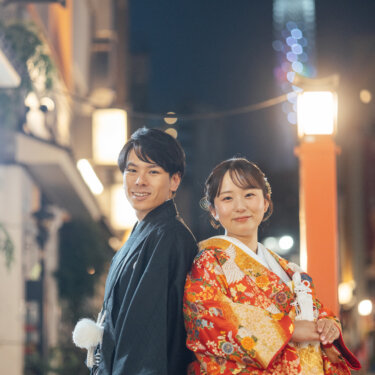 背中合わせで微笑む羽織袴の新郎と金糸の刺繍が映える色打掛の花嫁を、夜の浅草の街並みとスカイツリーを背景に撮った和装前撮り