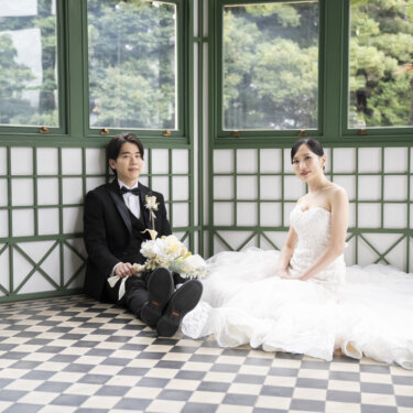 サンルームの壁にもたれて床に座り、ほほえみ合う新郎新婦。ドレスの裾が広がる旧古河邸でのナチュラルな前撮り写真