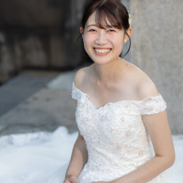 石段に座り、ビジューを散りばめたオフショルダーのウェディングドレスで満面の笑みを見せる花嫁。前撮りで残したいブライダルポートレート