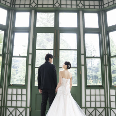 緑の格子窓が囲むサンルームで、窓の外を眺める新郎新婦の後ろ姿。洋館らしいクラシカルなウェディングフォト