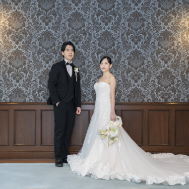 ブラケットライトが灯るダマスク柄の壁の前に並び、長いトレーンを広げたウェディングドレスの花嫁と新郎。旧古河邸で撮るレトロな前撮り