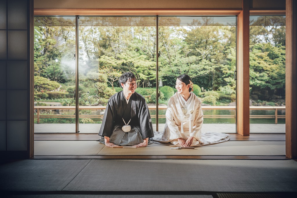 広縁から緑豊かな日本庭園を望む、黒紋付袴姿の新郎と白無垢姿の花嫁。開け放たれた障子越しの開放的な一枚。