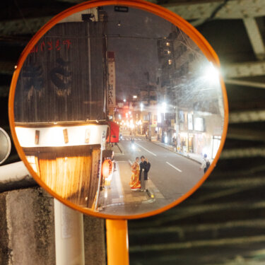 夜の路地のカーブミラーに小さく映り込む和装の新郎新婦。鏡越しに街並みを切り取った個性的な前撮り写真の構図