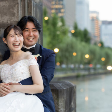 お濠沿いの並木を背景に、後ろからハグされて楽しそうに笑うオフショルダーのドレスの花嫁。自然な笑顔を引き出す洋装前撮り