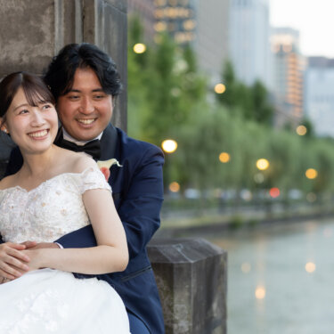 石の壁にもたれ、後ろから花嫁を抱きしめて笑うタキシードの新郎。お濠と並木のボケが美しい東京の前撮り