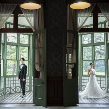 旧古河邸にて、左右の扉の奥にそれぞれ立つ新郎と花嫁を、シンメトリーに収めたサンルームの前撮り写真。構図が印象的な一枚