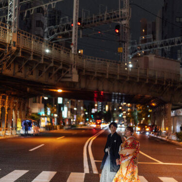高架の鉄橋が続く夜の横断歩道で寄り添う黒紋付袴の新郎と色打掛の花嫁。都会の街並みを背景にしたクールな和装前撮り