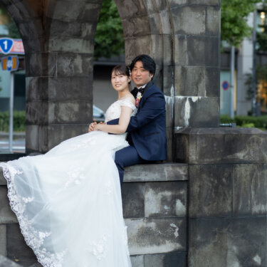 石造りのアーチの縁に並んで腰かけ、レースのトレーンを垂らして寄り添う新郎新婦。丸の内の街並みで撮るウェディングフォト