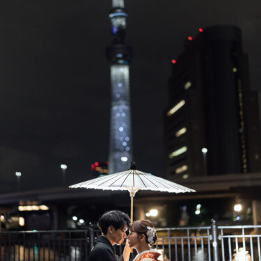 白い和傘をかざして川沿いの柵の前で顔を寄せ合う新郎新婦と、奥にそびえる夜の東京スカイツリー。浅草の夜景で撮る和装フォトウェディング