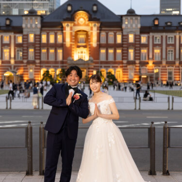灯りのともる東京駅の駅舎を背景に、胸の前で手を合わせて並ぶネイビーのタキシードの新郎と花嫁。おそろいのポーズがかわいい洋装前撮り