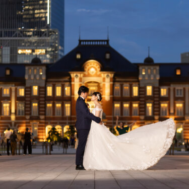 ブルーアワーの東京駅丸の内駅舎の前で、花嫁のドレスの裾をなびかせて寄り添う新郎新婦。赤レンガの駅舎が灯る洋装ロケーションフォト