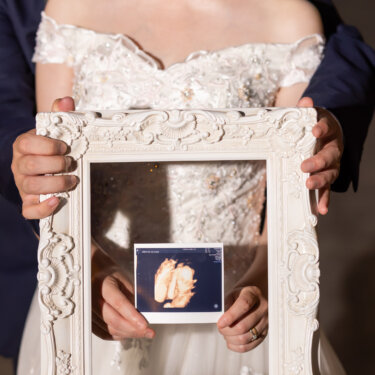 白いアンティークの額縁の中にエコー写真を掲げる新郎新婦の手元。赤ちゃんの報告にもなるマタニティのウェディングフォト