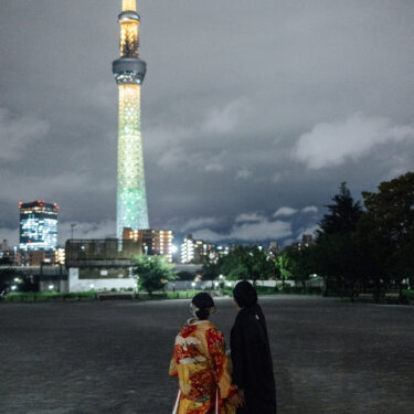 ライトアップされた東京スカイツリーを見上げて寄り添う、赤い色打掛の花嫁と黒紋付袴の新郎の後ろ姿。夜の浅草で撮る和装前撮りならではの一枚