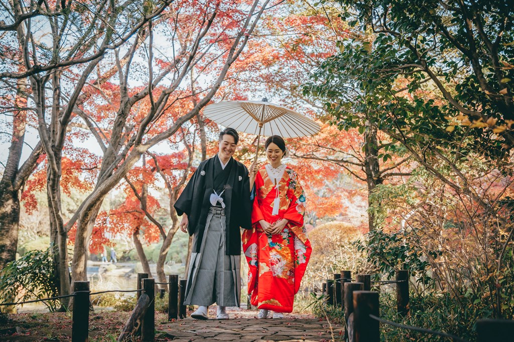 紅葉が色づく庭園の小道で、白い和傘をさして寄り添い歩く黒紋付袴姿の新郎と朱色の色打掛姿の花嫁。