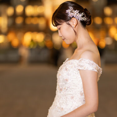 夜の東京駅の灯りをぼかした背景に、お腹にそっと手を添えてうつむく花嫁。花の髪飾りとオフショルダーのドレスが映えるマタニティ前撮りの横顔