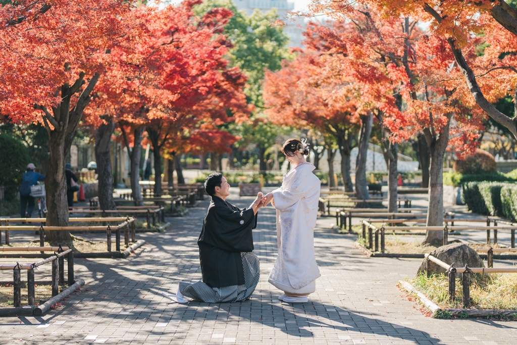 紅葉の並木道で片膝をつき、指輪を差し出す黒紋付袴姿の新郎と、感激した様子の白無垢姿の花嫁のプロポーズシーン。