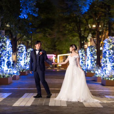 青いイルミネーションが輝く夜の並木道で、手をつないで向き合う新郎新婦。丸の内の街で撮るナイトフォトの洋装前撮り