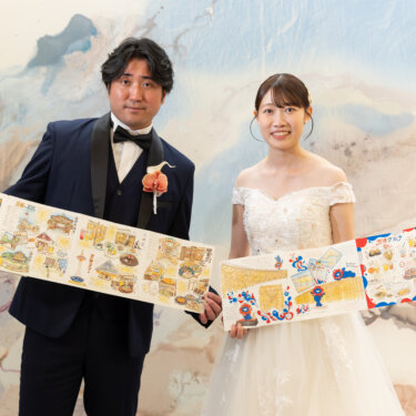 マーブル模様の壁の前で、手描きのイラストマップを広げて笑うタキシードの新郎とオフショルダーのウェディングドレスの花嫁。遊び心のある小物を使った前撮りの一枚