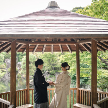 目白庭園の池のほとりにある六角の東屋で見つめ合う白無垢と紋付袴の新郎新婦。池と新緑を背景にした和装フォトウェディング