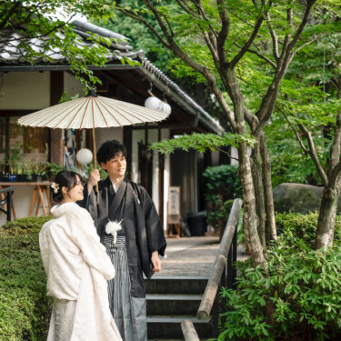 目白庭園の茶室の前で、白い和傘を差した紋付袴の新郎に寄り添って歩く白無垢の花嫁の和装前撮り