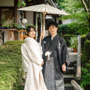 石段の上で白い和傘を差し、笑顔で並ぶ白無垢と紋付袴の新郎新婦。和傘を使った和装前撮りの定番カット