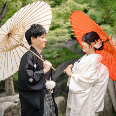岩と松のある日本庭園で、白い和傘の新郎と赤い和傘の花嫁が見つめ合うポーズ。色違いの和傘を使った和装前撮り