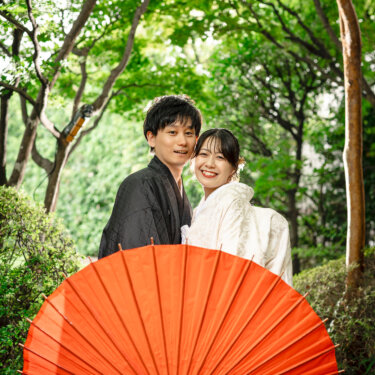 赤い番傘を手前に大きく開き、肩を寄せて笑う新郎新婦。目白庭園の木々を背景にした和装ロケーションフォトの構図