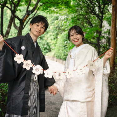 梅の花の形の『結婚しました』ガーランドを掲げて歩く白無垢と紋付袴の新郎新婦。結婚報告に使える和装前撮りのアイテムカット