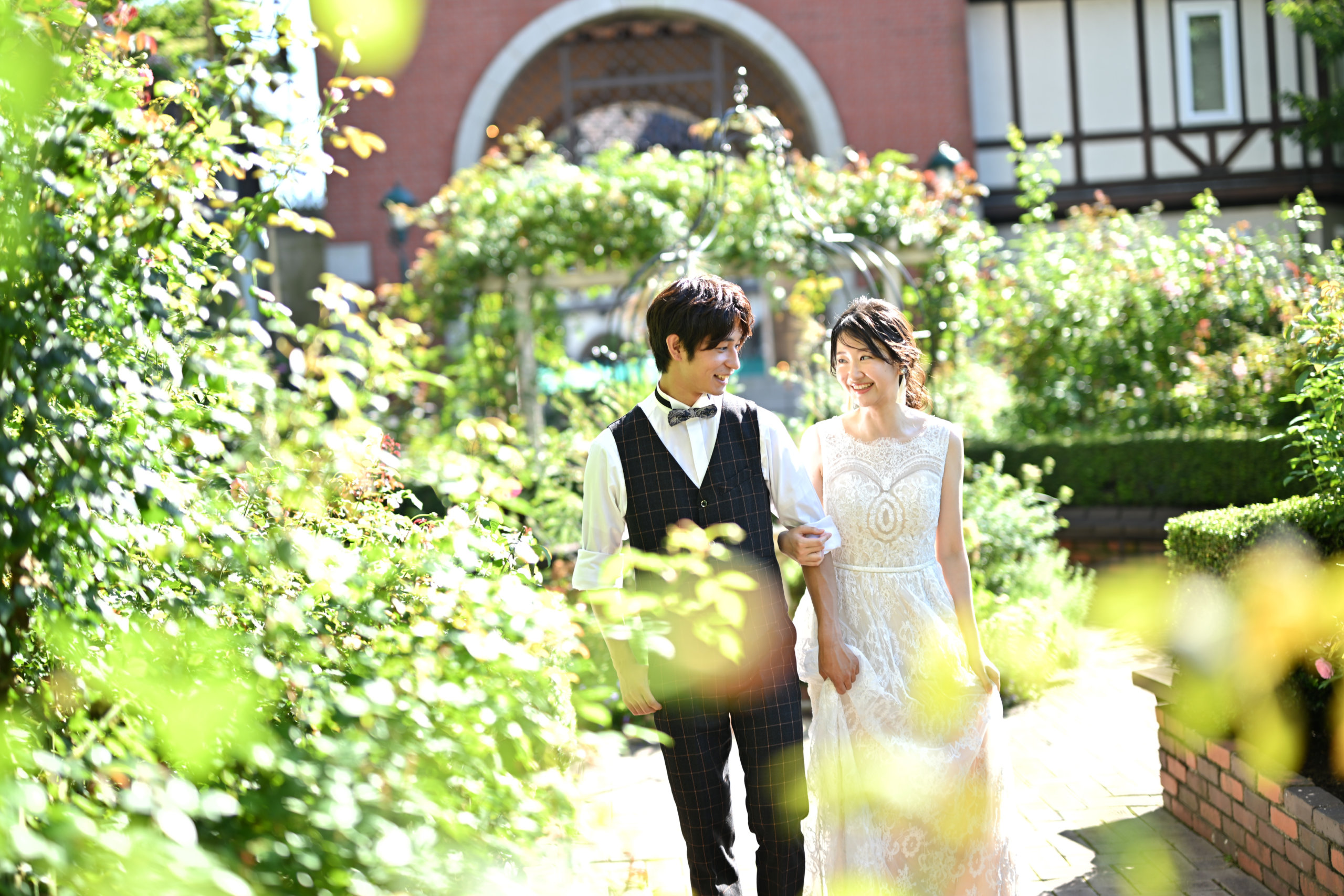 バラと緑が茂る洋館の庭を、腕を組んでゆっくり歩くレースのウェディングドレス姿の花嫁と新郎のロケーションフォト