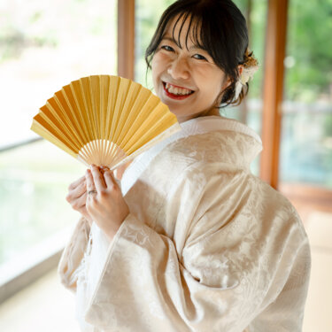 回廊の窓辺で金色の扇子を顔の横に広げて笑う白無垢のプレ花嫁。和装前撮りで人気の扇子ポーズ