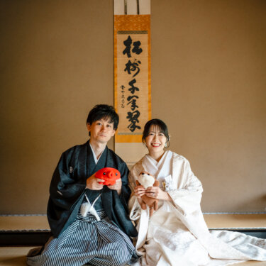 和室で正座し、赤い鯛と白いぬいぐるみを手に笑う新郎新婦。ぬいぐるみを小物に使ったかわいい和装前撮り