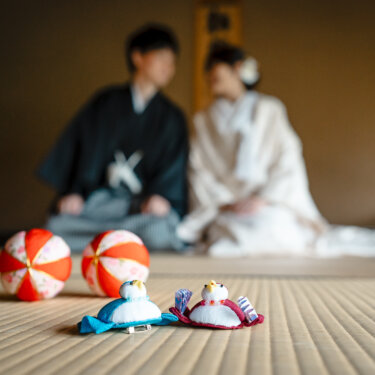 畳に置いた赤い手まりと鯛のぬいぐるみにピントを合わせ、奥で寄り添う新郎新婦をぼかした一枚。和室の和装前撮りで定番の小物カット