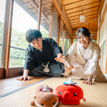 畳に手をついて笑いながらぬいぐるみに手を伸ばす新郎新婦。和室での和装前撮りに遊び心を加えたポーズ