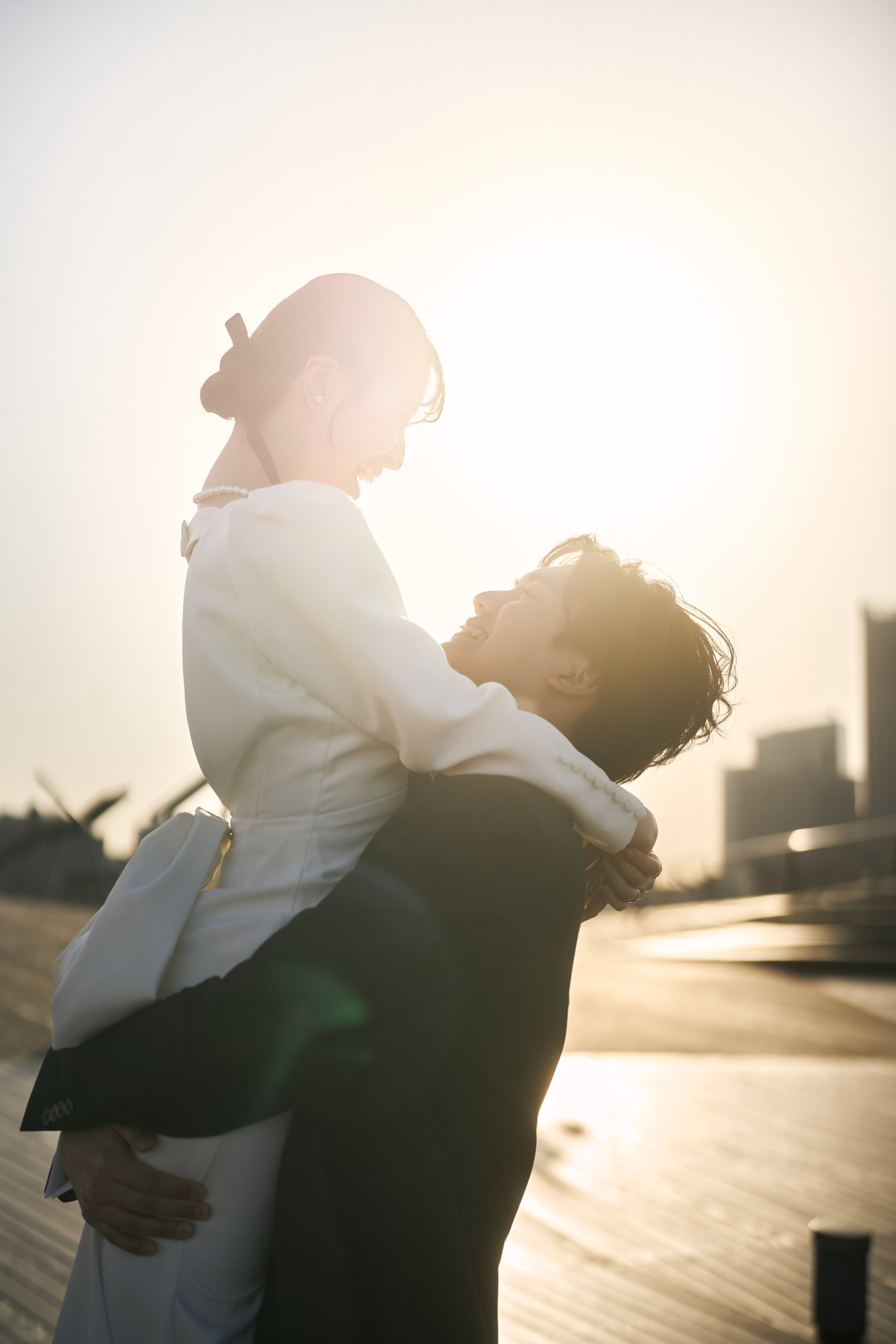 夕陽の逆光に包まれたウッドデッキで、新婦を抱き上げて笑い合う新郎新婦のサンセットウェディングフォト