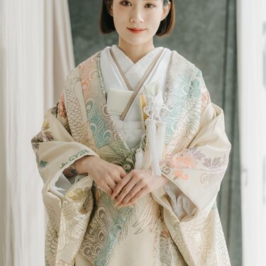 淡いクリーム地に孔雀と花模様を金糸で織り出した色打掛をまとい、手を重ねて微笑む花嫁。カーテンの光が差すスタジオでの和装前撮り