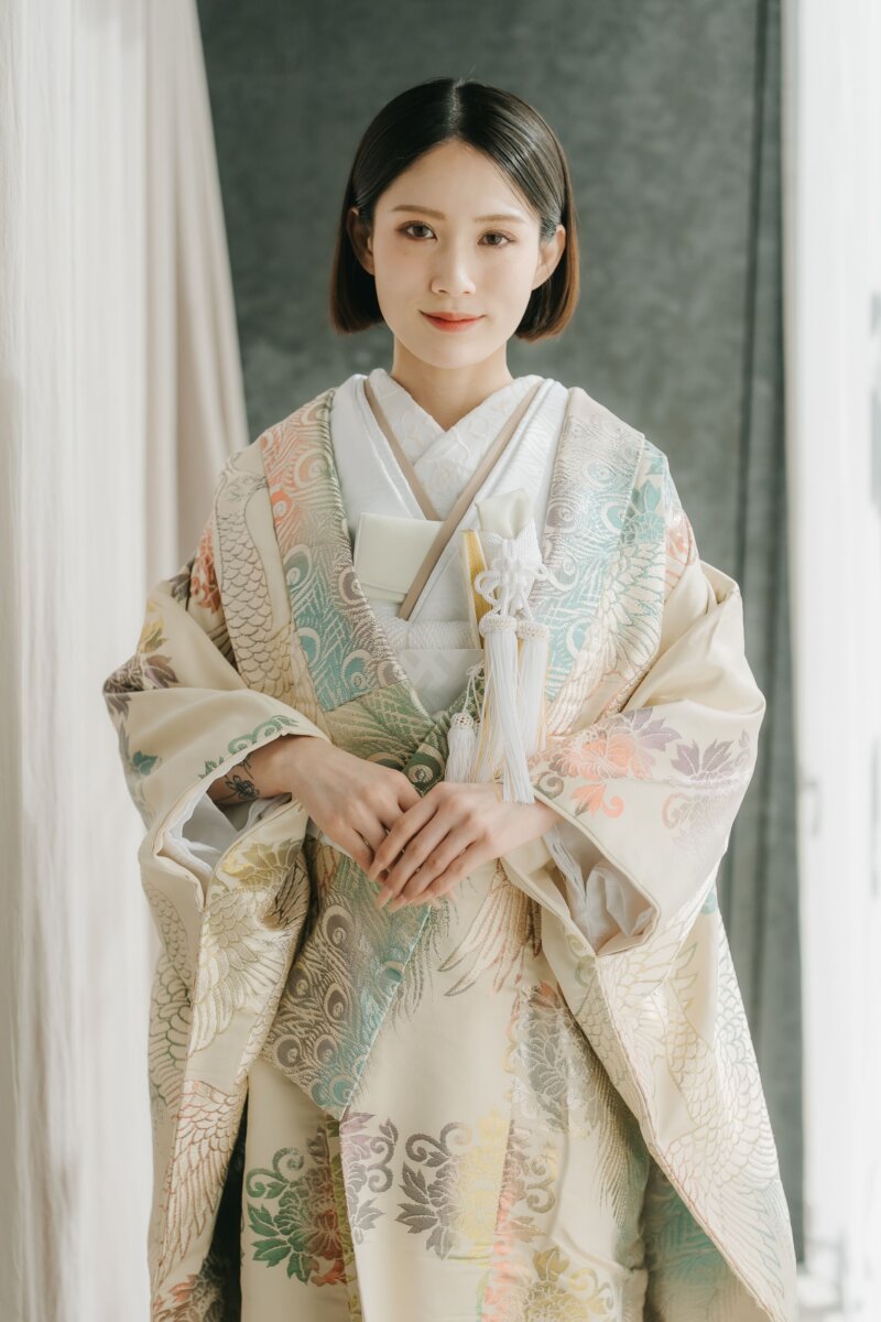淡いクリーム地に孔雀と花模様を金糸で織り出した色打掛をまとい、手を重ねて微笑む花嫁。カーテンの光が差すスタジオでの和装前撮り