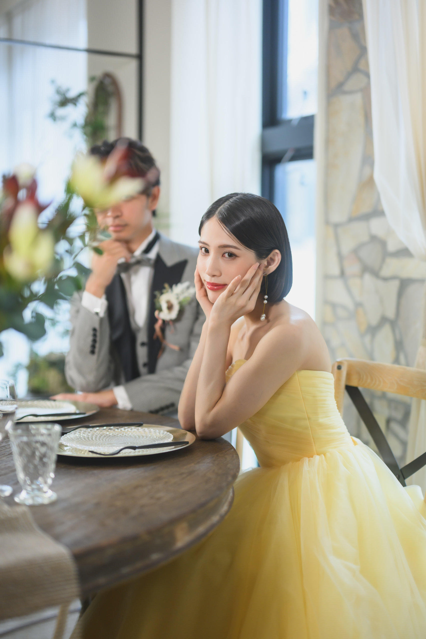 イエローのカラードレスの新婦とグレーのタキシード姿の新郎が、光の差し込む席に着く前撮りの一場面
