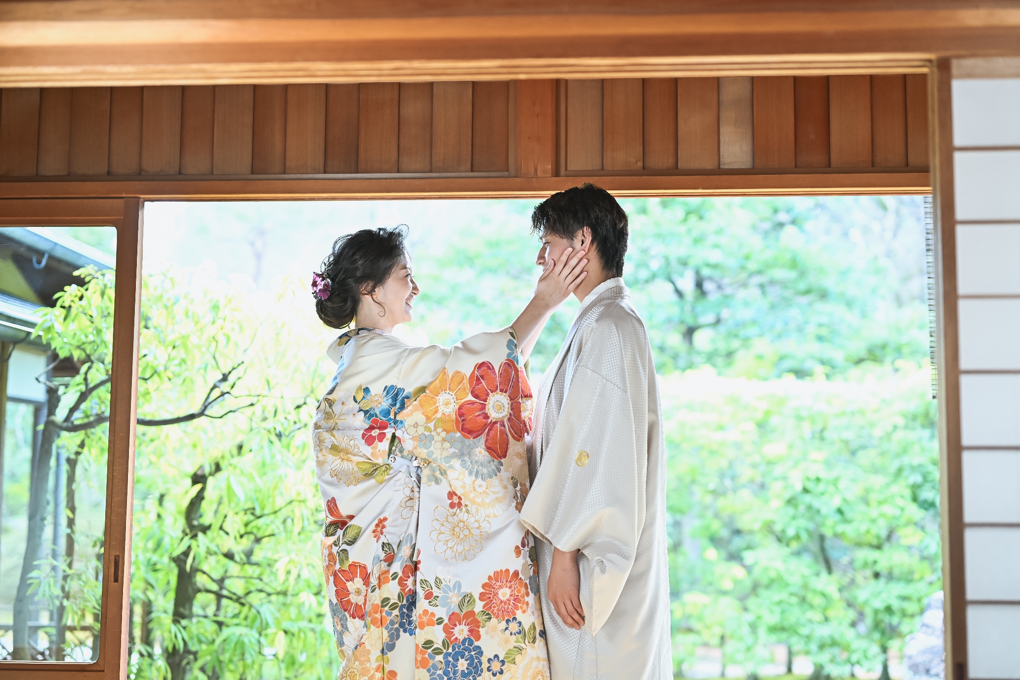緑あふれる庭に面した縁側で、色鮮やかな花柄の色打掛の花嫁が白い紋付姿の新郎の頬にそっと手を添える和装前撮り