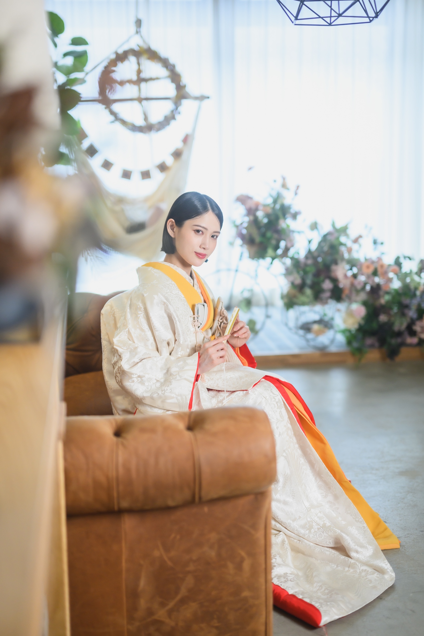ドライフラワーの装花に囲まれたスタジオで、白無垢に橙の差し色をまとい末広を手にソファへ腰かける花嫁の和装前撮り（東京駅店）
