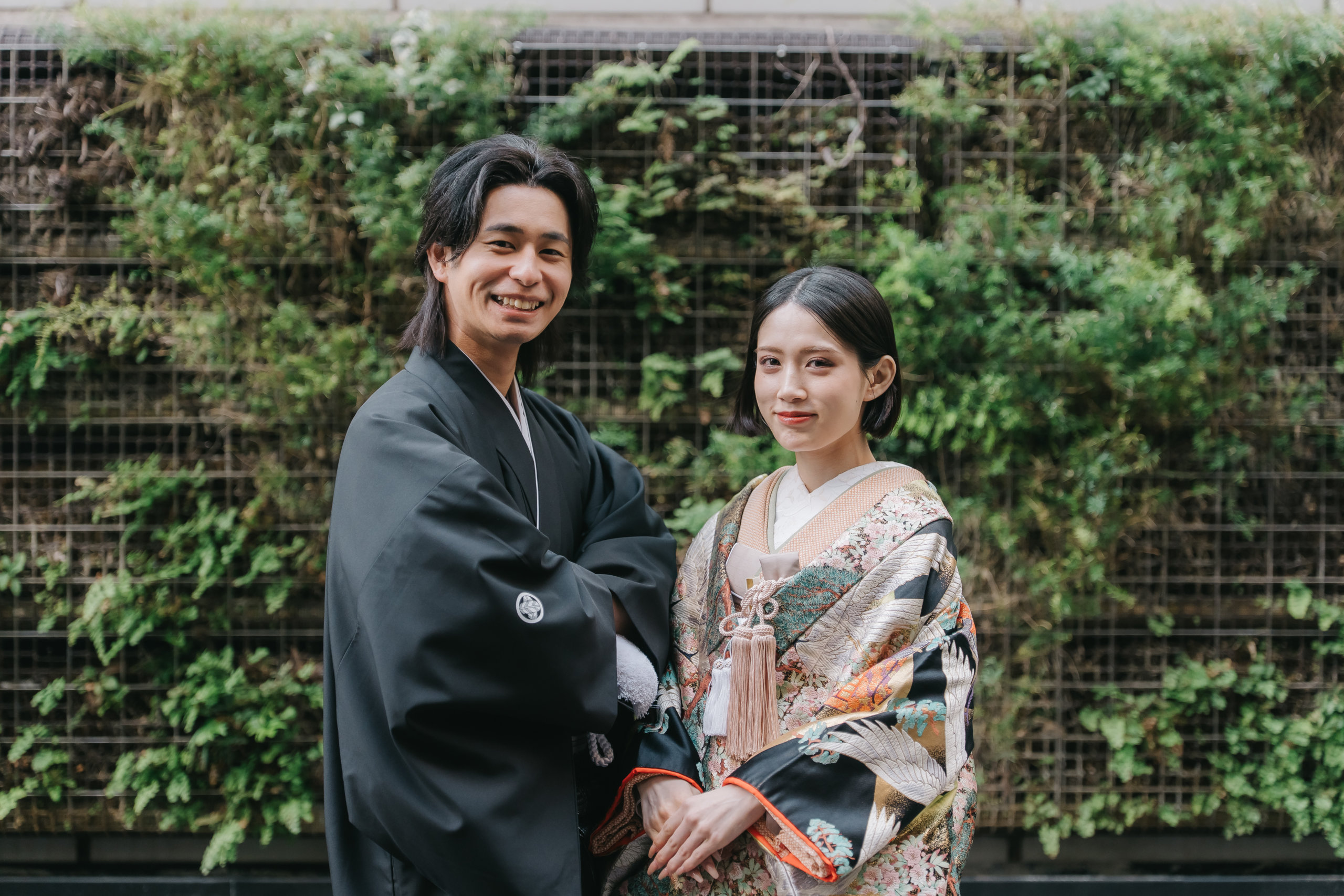 一面のグリーンウォールを背に、鶴柄の色打掛の花嫁と黒紋付羽織袴の新郎が並んで微笑む和装フォトウェディング