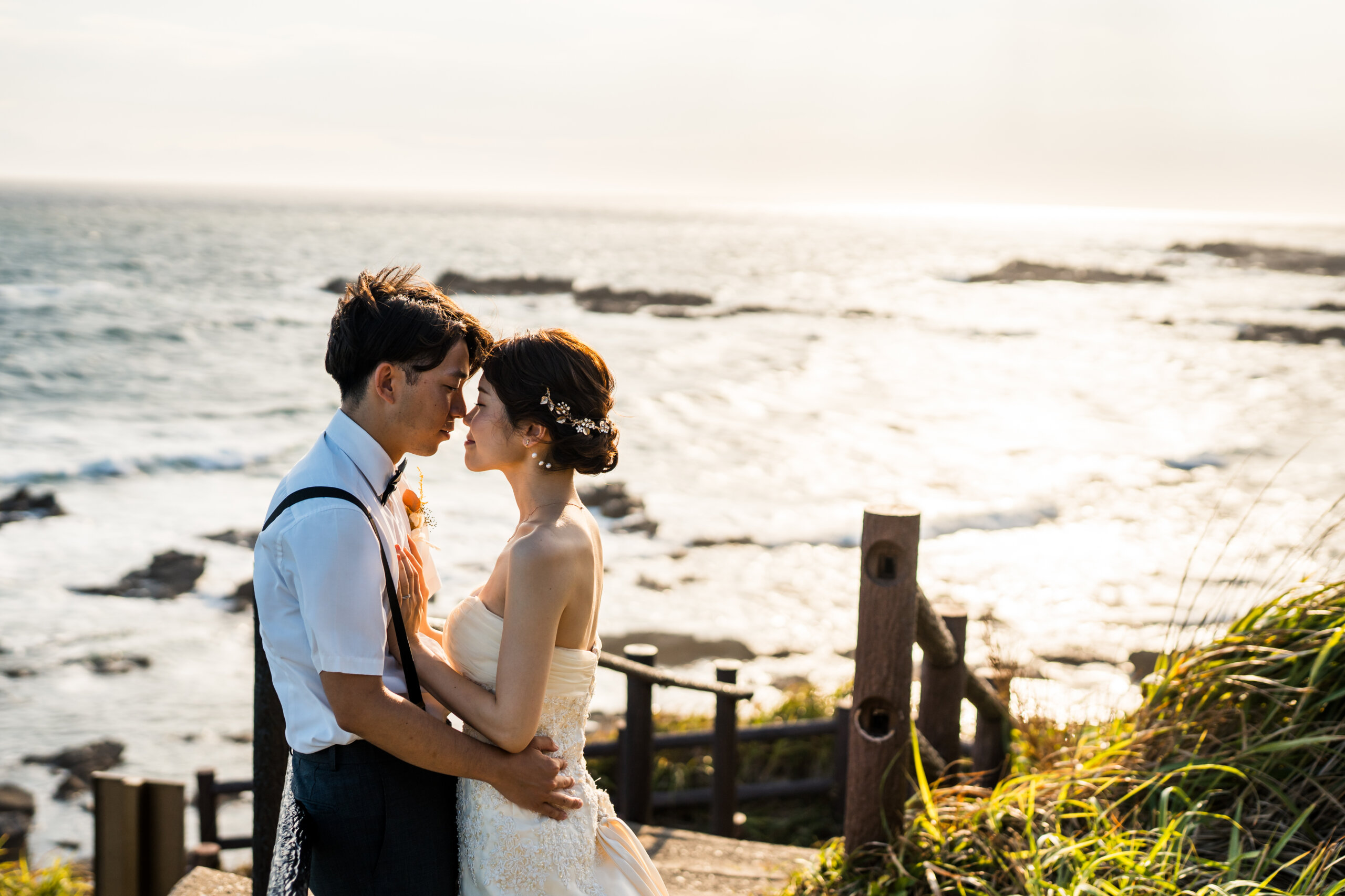 城ヶ島の海を見渡す展望スポットで寄り添うウェディングドレスの花嫁とタキシードの新郎。海と空が広がるロケーションフォト