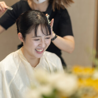 ヘアメイク中に笑顔を見せる花嫁と、髪を整えるスタッフ。和装前撮り当日の支度風景を残したヘアアレンジのひとこま
