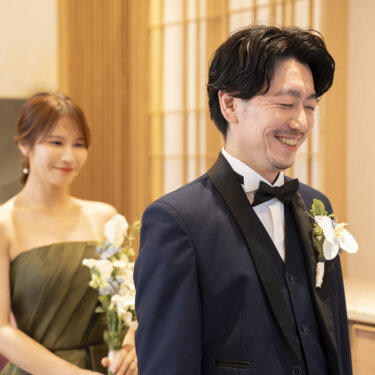 ブーケを持ってそっと近づく花嫁に気づかず微笑む、蝶ネクタイの新郎のアップ。東京駅店のスタジオで撮るファーストミートの前撮り