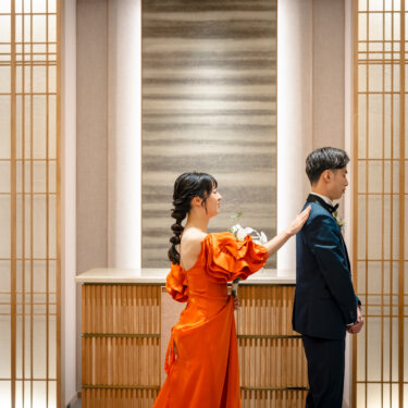 木の格子が並ぶチャペルで、新郎の肩にそっと触れて振り向かせるオレンジのカラードレスの花嫁。ファーストミートを再現した前撮りのワンシーン