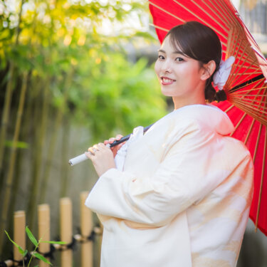 赤い和傘を肩にさして振り返り、竹林を背に微笑む白無垢の花嫁のソロカット。結婚写真に映える和装ポーズ