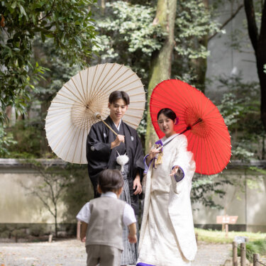 白と赤の和傘を差した紋付袴の新郎と白無垢の花嫁へ、石畳を歩いて向かう男の子。小石川後楽園の和装前撮り
