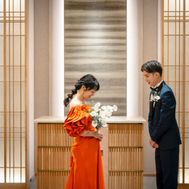 和モダンなチャペルの祭壇前で、手元のブーケを見下ろすカラードレスの花嫁と、隣で微笑むタキシードの新郎を撮った前撮りの一枚