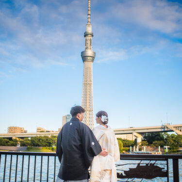 隅田川の手すり越しに東京スカイツリーを見上げる紋付袴の新郎と白無垢の花嫁の後ろ姿。浅草の和装前撮り
